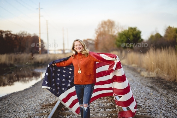 Smiling female holding the United States flag while walking on the train rails
