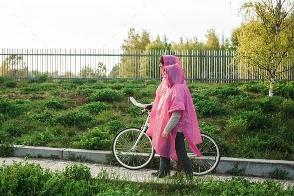 Male and a female in pink plastic raincoats walking through the road with a bicycle on a date