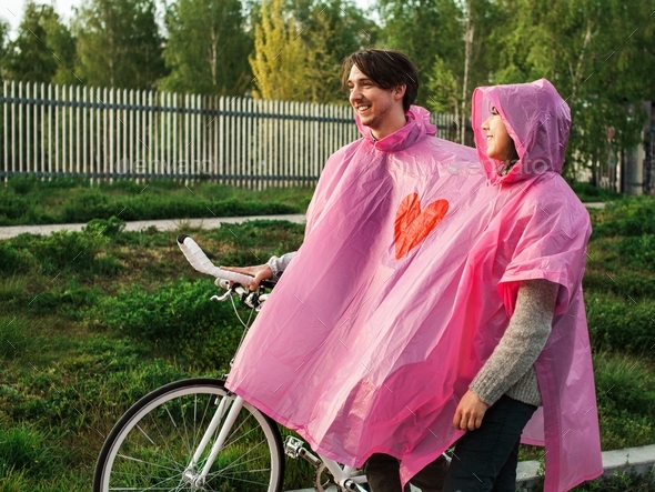 Male and a female in a shared pink plastic raincoat walking with a bicycle on a date