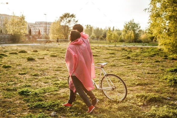 Male and a female in pink plastic raincoats walking in the field with a bicycle on a date