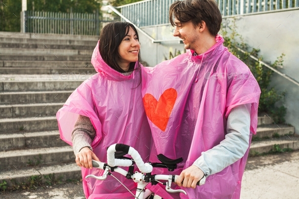 Closeup shot of a male and a female sharing a pink plastic raincoat with a red heart in the center