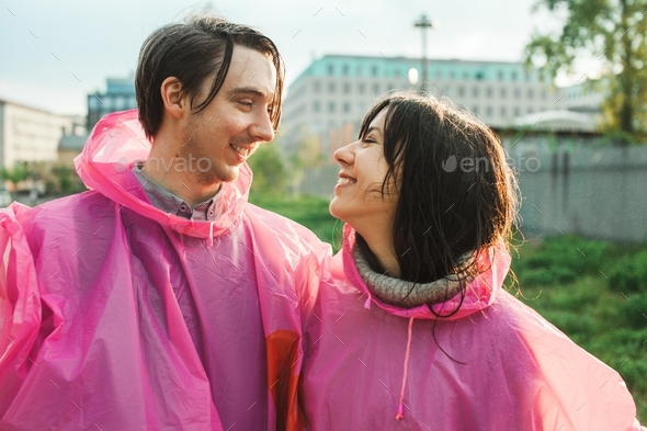 Closeup shot of a male and a female in pink plastic raincoats smiling at each other romantically
