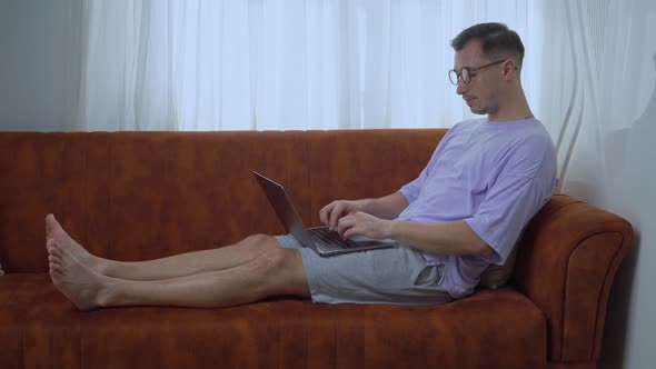 A Man with Glasses Typing on a Computer Working From Home on the Couch