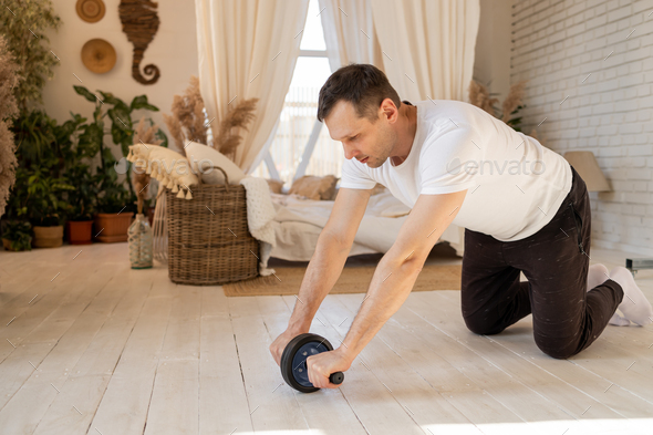 Athletic man is doing exercises with an abs wheel in living room at home. Sport and fitness. Workout