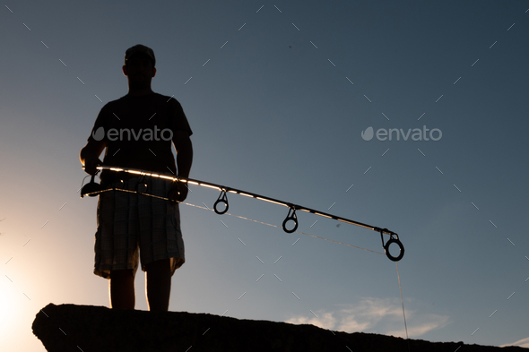 Low angle shot of the silhouette of a fisherman standing on the rocks with  a rod Stock Photo by wirestock