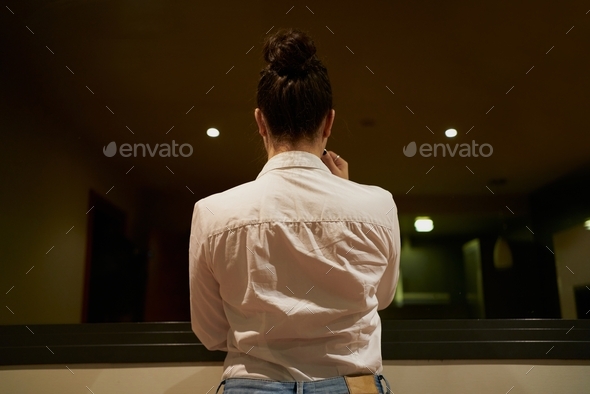 Closeup of the back profile of a woman wearing white blouse looking through a hotel window at night