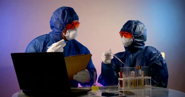 Two People in Protective Clothing, Respirators, Glasses and Gloves Are Sitting at a Table. They alt