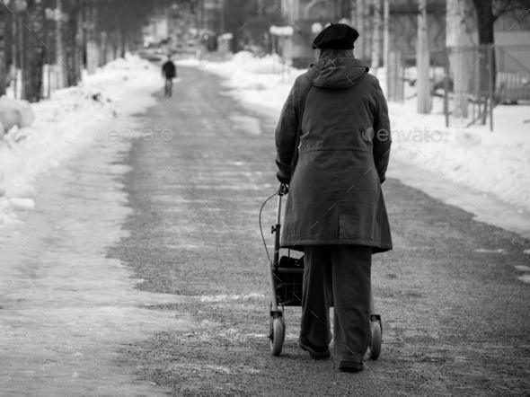 Grayscale shot of an elderly woman walking with a walker down the snow-covered street