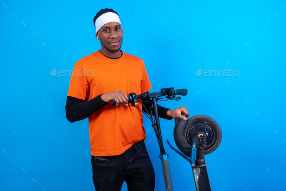 Portrait of a black ethnic man in orange clothes on a blue background, with an electric skateboard