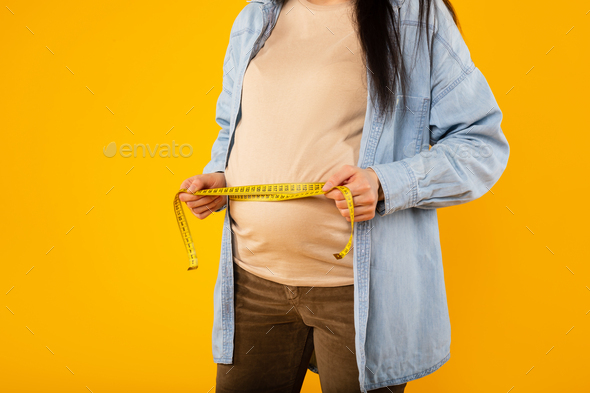 Young pregnant lady measuring her belly with tape to keep track of her fetus development, cropped