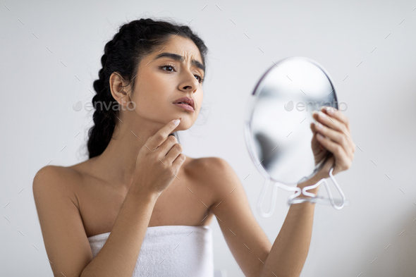 Worried Indian Female Holding Magnifying Mirror And Looking At Pimple On Chin