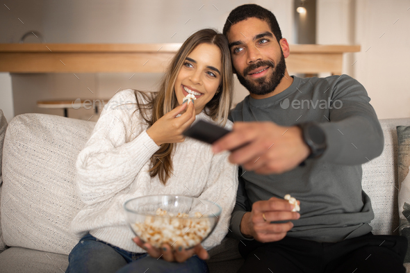 Cheerful young arab guy with beard and european lady watch tv with remote control, eat popcorn