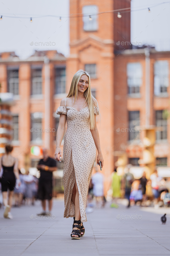 Vertical shoot of walking blonde young Swedish Woman in beige dress walks  by street over blurry city Stock Photo by ionadidishvili