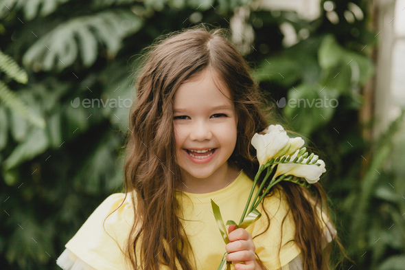 Cute little girl in a yellow dress, holding spring flowers in her hands, standing against  - Stock Photo - Images