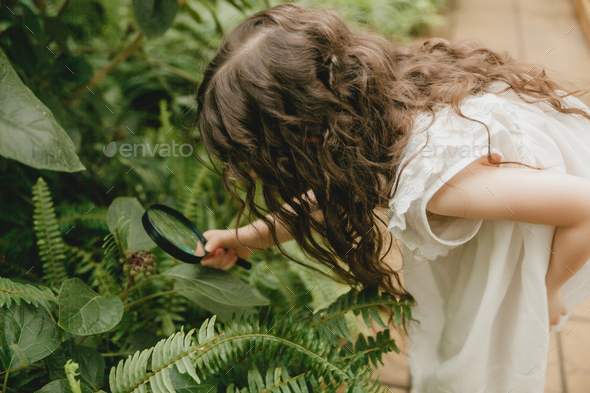 Portrait of a cute little girl looking at plants through a magnifying glass.  - Stock Photo - Images