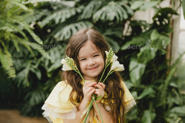 Cute little girl in a yellow dress, holding spring flowers in her hands, standing against - Stock Photo - Images