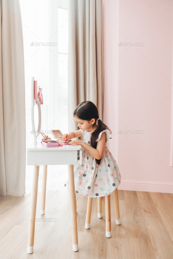 a beautiful little girl in the mirror preening. a little girl is sitting at a children's table - Stock Photo - Images