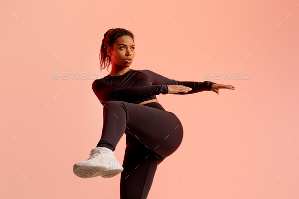 Fit african american lady in sportswear warming up and lifting leg up, exercising on peach neon