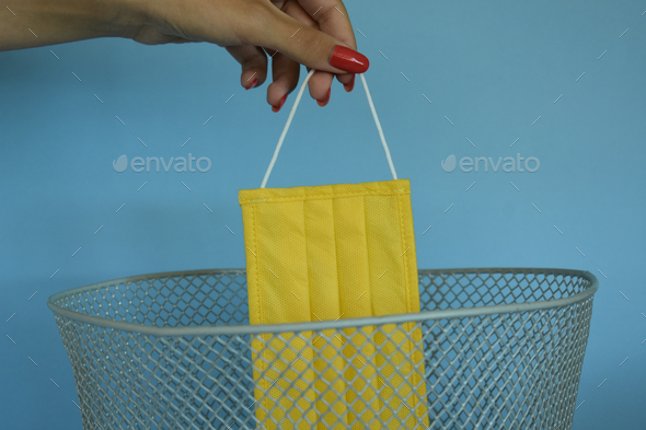 Female hand throwing a new yellow medical face mask into a trash can, blue background