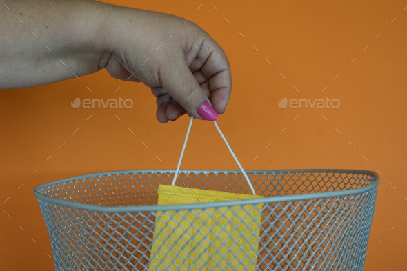Female hand throwing a new yellow medical face mask into a trash can, orange background