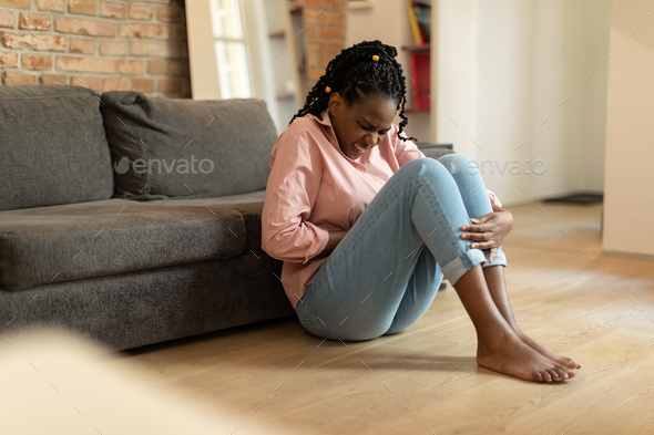 Young african american woman suffering from stomach ache, touching tummy, sitting on floor near sofa
