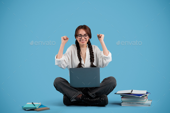 Cheerful european teen girl student with pigtails in glasses sits on floor with books and pc