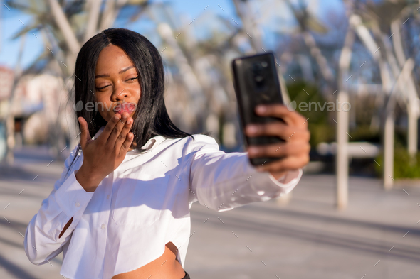 Young african woman in white clothes in the city at sunset, video call, blowing a kiss
