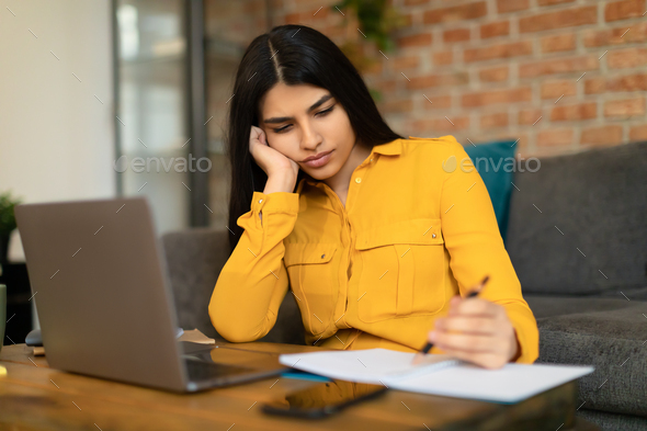 Bored spanish teen girl studying online from home, taking notes during online lesson, participating