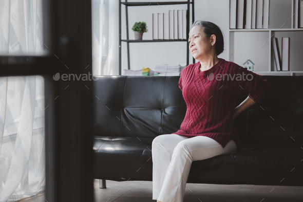 Senior woman sitting in the living room at home and showing body aches, aging sickness, body aches o
