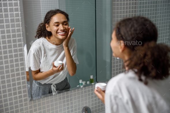 Smiling woman applying anti-wrinkle cream standing behind mirror in home bathroom. Beauty procedure
