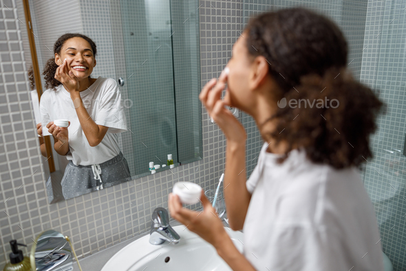 Smiling woman applying anti-wrinkle cream standing behind mirror in home bathroom. Beauty procedure