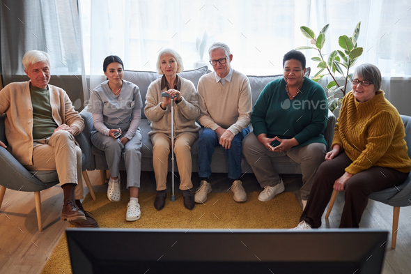 Elderly people watching TV in retirement home sitting in row on sofa