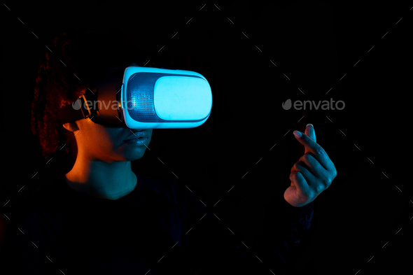 Black woman wearing virtual reality goggles and making heart sign with hand  in black light, studio Stock Photo by Prostock-studio