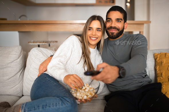 Smiling millennial arabic guy with beard and remote control hugs european lady, watch TV, eat