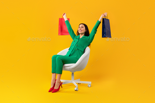 Full body length shot of contented shopaholic woman sitting on chair and raising hands with shopping - Stock Photo - Images