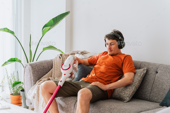 Tired man holding wireless vacuum cleaner relaxing chilling on the couch surrounded with plants