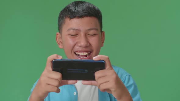 Laughing Young Asian Boy Looking At The Phone Screen In Green Screen In The Studio alt