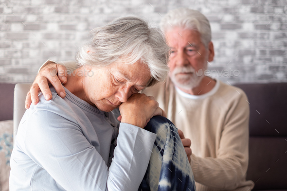 Sad and depressed elderly woman sitting at home while her husband tries to comfort her