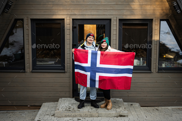 Portrait of couple outside cabin house holding Norway flag. Scandinavian culture, norwegian people.