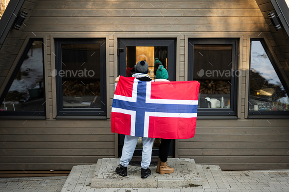 Back of couple outside cabin house holding Norway flag. Scandinavian culture, norwegian people.