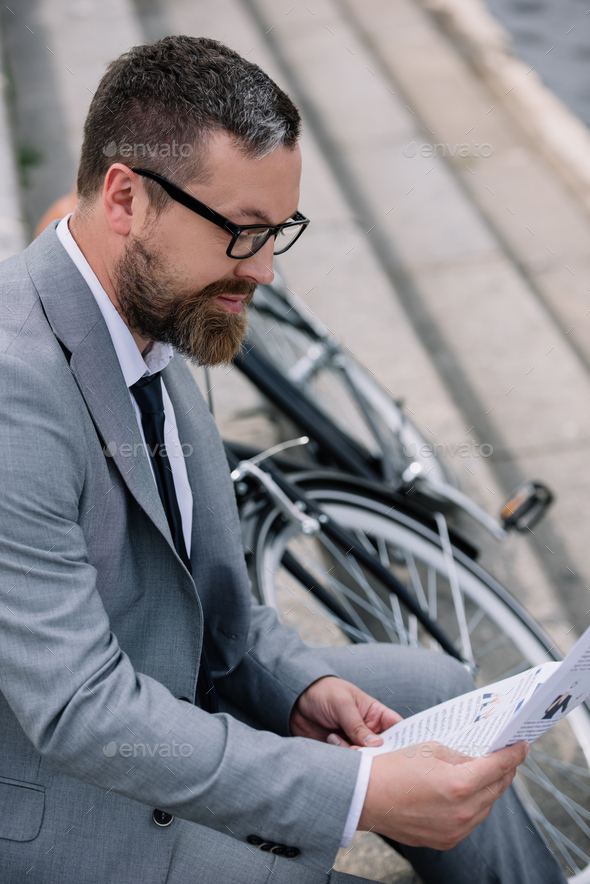 handsome bearded businessman reading newspaper on stairs with bicycle