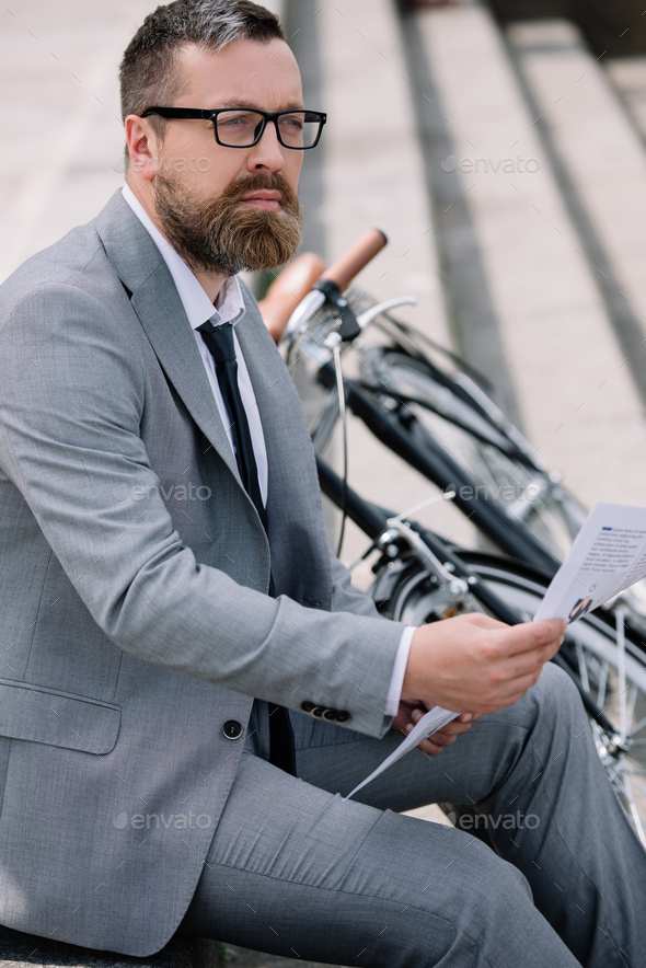 pensive bearded businessman reading newspaper on stairs with bicycle