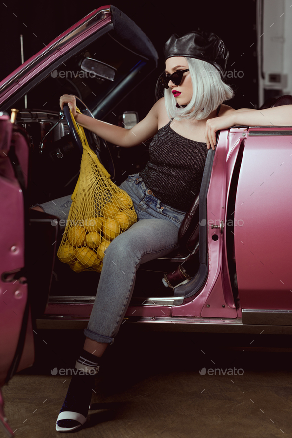 beautiful stylish blonde girl in beret and sunglasses holding string bag with lemons while sitting
