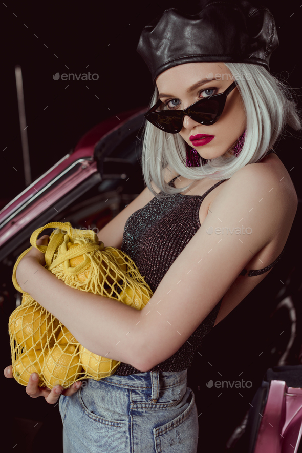 beautiful blonde woman in sunglasses and beret holding string bag with lemons and looking at camera