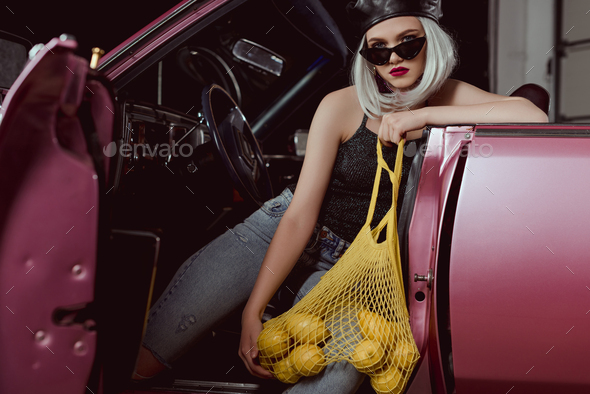 stylish blonde girl in beret and sunglasses holding string bag with lemons while sitting in car