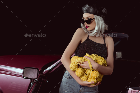 beautiful blonde woman in sunglasses and beret holding string bag with lemons and looking away while