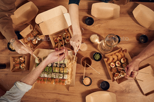 Top view of table with sushi on it in paper eco boxes, hands of the people taking it, eating
