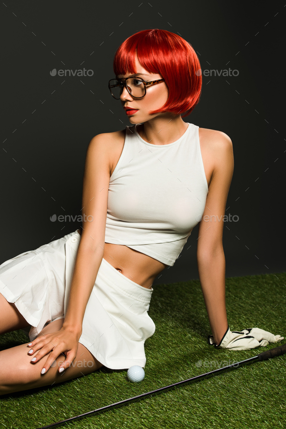 seductive young woman with red bob cut sitting on green grass with golf equipment on grey