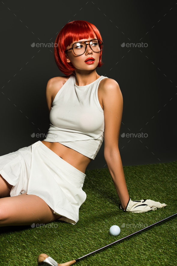 attractive young woman with red bob cut sitting on green grass with golf equipment on grey