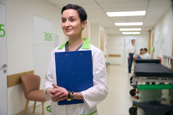 Woman in a medical gown stands with folder in corridor
